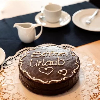 Eine Schokoladentorte mit der Aufschrift „Schönen Urlaub“ steht auf einem Tisch. Darum liegen Tassen und Untertassen, bereit für eine Kaffeerunde.