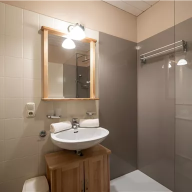 Ein modernes Badezimmer mit einem Waschbecken, einem Spiegel und einer Relaxdusche. Die Wände sind hell und sauber, mit einem Holzregal für Handtücher.