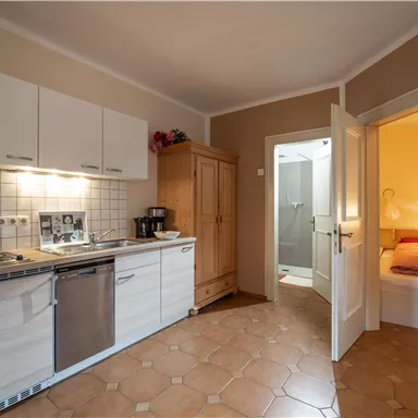 Eine moderne Küche mit hellen Schränken und einem Kochfeld. Im Hintergrund befindet sich ein Zugang zu einem Badezimmer und einem Schlafzimmer.