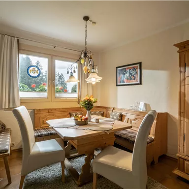 Ein gemütlicher Essbereich mit einem Holztisch und weißen Stühlen. Durch das Fenster ist Blick auf einen Garten mit Blumen sichtbar.