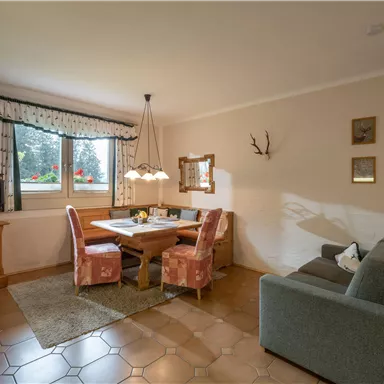 Ein gemütlicher Essbereich mit einem Holztisch und bunten Stühlen. Die Wand ist mit Bildern und Jagdtrophäen dekoriert, und ein Fernseher steht in der Ecke.