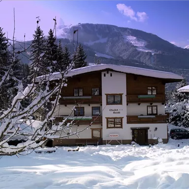 Ein gemütliches Chalet im Schnee, umgeben von schneebedeckten Bäumen. Im Hintergrund sind majestätische Berge und ein klarer Himmel zu sehen.