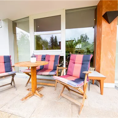 Ein gemütlicher Außenbereich mit Holzstühlen und einem kleinen Tisch. Die Sitzgelegenheiten sind in hellen Farben und laden zum Entspannen ein.