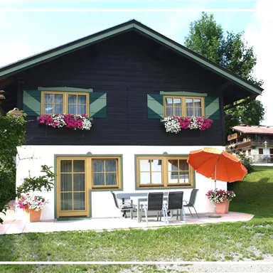 Ein charmantes, zweigeschossiges Holzhaus mit bunten Blumenfenstern. Vor dem Haus steht ein Tisch mit Stühlen und einem roten Sonnenschirm.