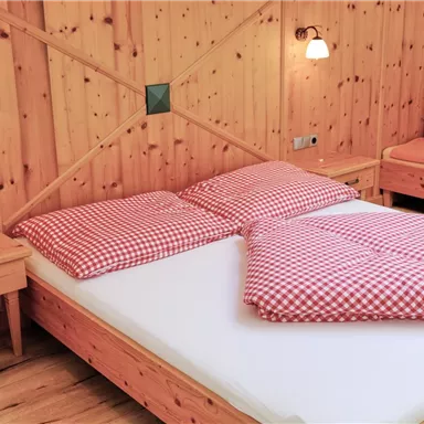 Ein gemütliches Schlafzimmer mit einem Holzbett und karierten Kissen. Die Wände sind mit Holz vertäfelt, und es gibt eine bequeme Sitzgelegenheit im Hintergrund.