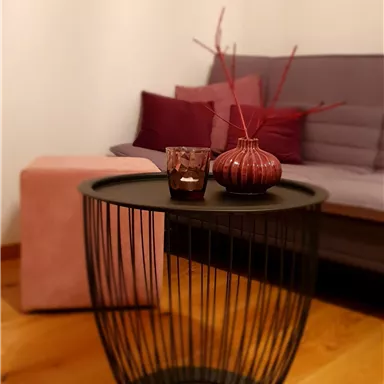 A modern side table with a wire frame is in a cozy living room. On the table, there is a candle and a decorative vase with branches placed.