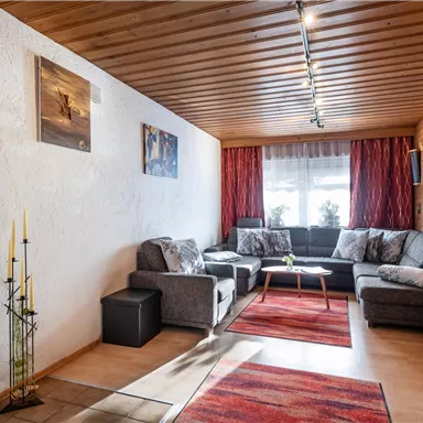 A cozy living room with gray sofas and a wooden floor. There are light walls, pictures on the wall, and a window with a red curtain.