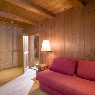 A cozy room with wooden-paneled walls and a red sofa. A lamp provides warm light, while an open room with doors in the background can be seen.