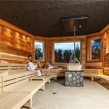 A modern wellness area with a wooden sauna and large windows that offer a view of nature. Two people relax on the benches and enjoy the calm atmosphere.
