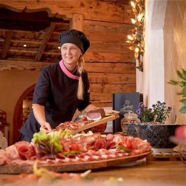 A cook presents a beautiful selection of sausages and vegetables on a table. In the background, rustic wooden walls and plants are visible.
