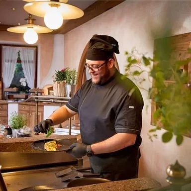 A cook is preparing a meal in a cozy kitchen. The environment is bright and friendly, with plants and decorative elements.