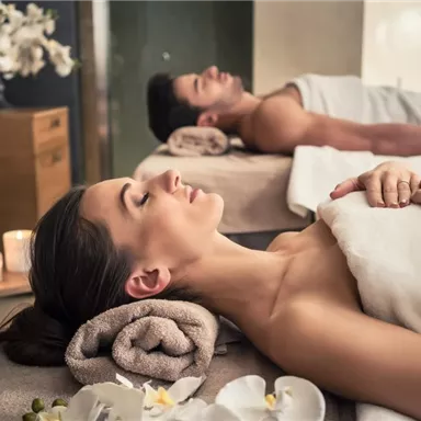 A relaxing spa experience with a couple lying on massage tables. Soft light from candles and flowers creates a calm atmosphere.