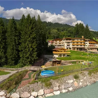 A picturesque hotel by the banks of a river, surrounded by green forests and mountains. The outdoor area offers playground equipment and a hot tub for guests.