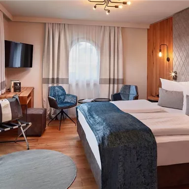 A modern hotel room with a large bed and stylish furniture. The windows are decorated with light curtains and a television is mounted on the wall.