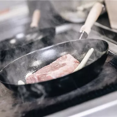 Ein Stück Fleisch brät in einer Pfanne auf dem Herd. Dampf steigt auf und es sind Küchenutensilien im Hintergrund zu sehen.
