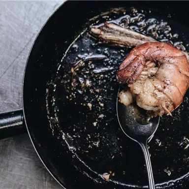 Eine Pfanne mit einem großen, gebratenen Garnelenstück und etwas Bratensatz. Im Hintergrund sind Reste von Kräutern zu sehen.