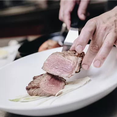 Ein Koch schneidet ein Stück saftiges Fleisch auf einem weißen Teller. Das Gericht wird liebevoll angerichtet und zeigt frische Beilagen.