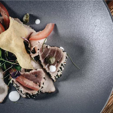 Ein kunstvoll angerichteter Teller mit dünn geschnittenem Fisch, garniert mit frischen Kräutern und Gemüse. Die Präsentation erfolgt auf einem dunklen, runden Teller.