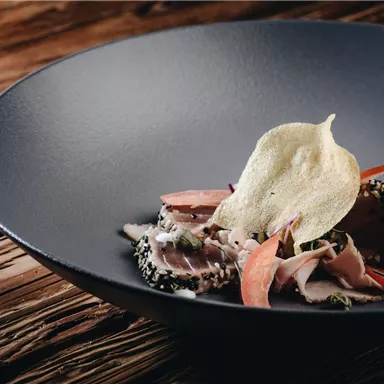 Ein ansprechend angerichtetes Gericht auf einem schwarzen Teller. Es enthält zarte Fleischstücke, Gemüse und eine knusprige Dekoration.