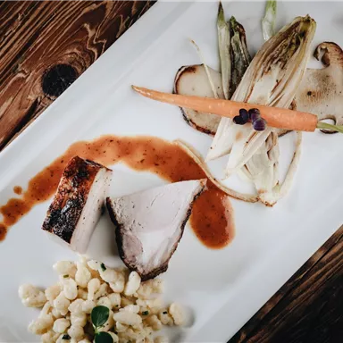 Ein elegant angerichteter Teller mit gegrilltem Fleisch, Gemüse und einer Sauce. Dazu gibt es eine Beilage aus kleinen Teigstücken.