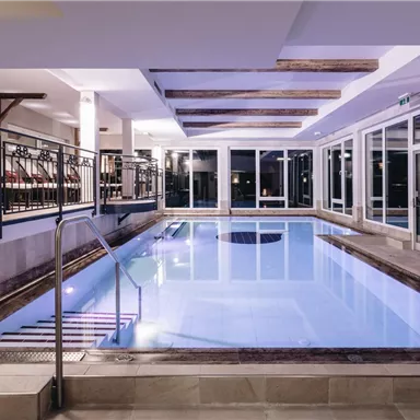 Ein modernes Hallenbad mit klarem Wasser und gemütlichen Sitzgelegenheiten. Große Fenster sorgen für eine helle Atmosphäre.