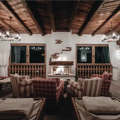 Ein gemütlicher Wohnraum mit Holzdecken und einem offenen Kamin. Bequeme Sessel und Fenster sorgen für eine einladende Atmosphäre.