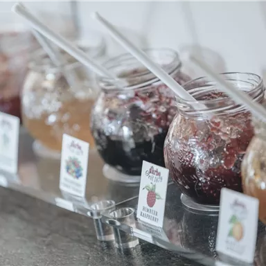 Bunte Gläser mit verschiedenen Marmeladen stehen auf einer Theke. Jedes Glas hat einen Löffel und ein kleines Schild mit der Geschmacksrichtung.