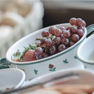 Ein schöner Obstteller mit Trauben und Erdbeeren in einer Schüssel. Im Hintergrund sind weitere Früchte und Brötchen sichtbar.