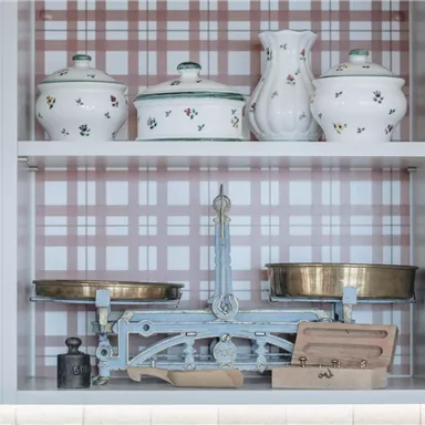 Eine dekorative Vitrine mit verschiedenen Küchenutensilien, darunter Töpfe und eine alte Waage. Der Hintergrund ist mit einem karierten Muster in sanften Farben gestaltet.