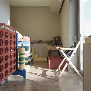 Ein gemütlicher Spielbereich mit bunten Spielzeugen und einem Stuhl. Der Raum hat große Fenster und viel Tageslicht.