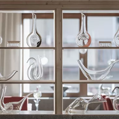 Eine Glaskunst-Vitrine mit verschiedenen dekorativen Glasobjekten. Die Objekte variieren in Form und Größe und sind vor einem hellen Hintergrund aus Fenstern arrangiert.