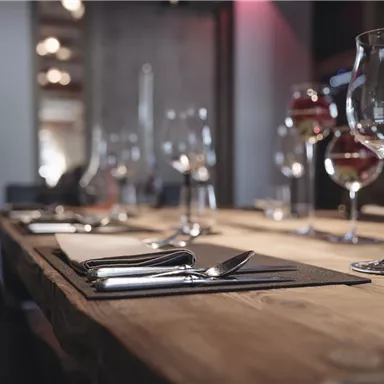 Ein stilvoll gedeckter Tisch in einem eleganten Restaurant. Es sind Gläser und Besteck auf einem Holzuntergrund platziert.