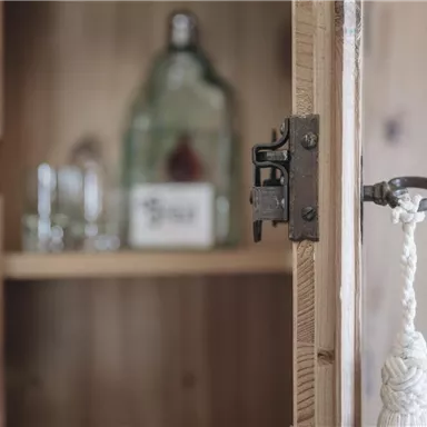 Ein geöffneter Holzschrank mit einem alten Schloss. Im Inneren sind ein paar Gläser und eine Flasche sichtbar.