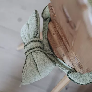 Ein Stuhl mit einem mintgrünen Kissen, das mit einer Schleife gebunden ist. Der Holzstuhl hat eine natürliche, warme Ausstrahlung.