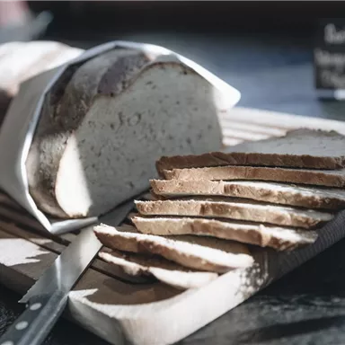 Ein frisch gebackenes Brot liegt auf einem Holzbrett, das teilweise in Scheiben geschnitten ist. Daneben liegt ein Messer und im Hintergrund ist ein Schild zu sehen.