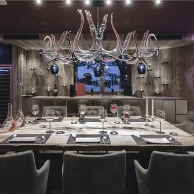 Ein stilvoll eingerichteter Speiseraum mit einem großen Esstisch und bequemen Stühlen. Im Hintergrund sind moderne Lichtelemente und eine Wand mit Weinflaschen zu sehen.