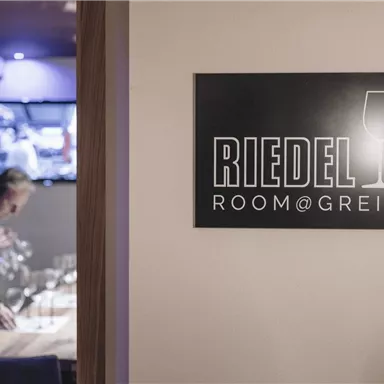 Ein Schild mit der Aufschrift "RIEDEL ROOM@GREIL" in einem eleganten Restaurant. Im Hintergrund ist eine Person, die einen Tisch vorbereitet.