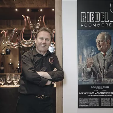 Ein Mann steht an einer Wand, die ein Porträt von Claus Josef Riedel zeigt, dem Erfinder des modernen Weinglases. Im Hintergrund sind Gläser und Weinaccessoires zu sehen.
