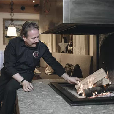 Ein Koch füttert das Feuer in einem gemütlichen Restaurant. Im Hintergrund sind bequeme Sitzgelegenheiten und eine angenehme Atmosphäre zu sehen.