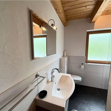 A modern bathroom with wooden ceilings and a large mirror. The walls are light and the floor is tiled in dark colors.