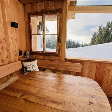Ein gemütlicher, holzverkleideter Raum mit einem runden Tisch. Draußen ist ein verschneites Landschaftsbild mit Bäumen im Hintergrund.