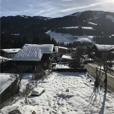 Eine verschneite Landschaft mit traditionellen Chalets und einem klaren blauen Himmel. Im Hintergrund sind die schneebedeckten Berge zu sehen.