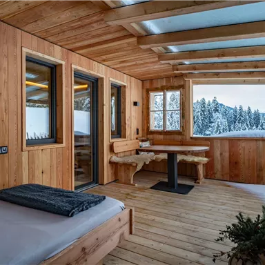 Eine gemütliche Veranda aus Holz mit einer Aussicht auf verschneite Bäume. Der Raum ist hell und einladend, ideal zum Entspannen.