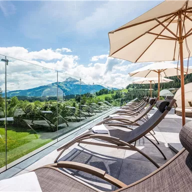 A modern terrace with loungers and sun umbrellas, surrounded by a beautiful mountain landscape. The sky is clear, and the atmosphere is relaxing.