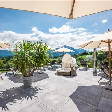 A stylish terrace with loungers and sun umbrellas. In the background, green mountains and a blue sky can be seen.