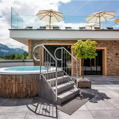 A modern terrace with a whirlpool and sun umbrellas. In the background, mountains and a clear sky are visible.