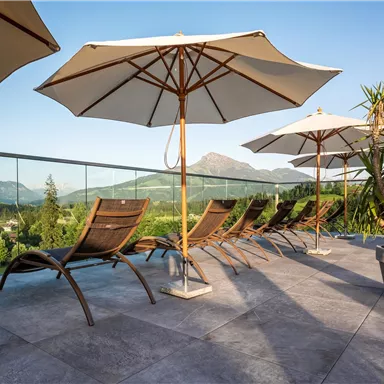 A relaxing area with lounge chairs and sun umbrellas on a terrace. In the background, mountains and a clear sky can be seen.