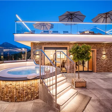 A modern house with a terrace and a hot tub. The surroundings are lit in the evening and offer a view of the mountains.