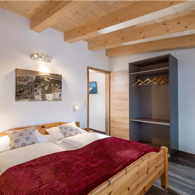 Ein gemütliches Schlafzimmer mit einem Doppelbett und roten Decken. Es gibt Holzdecken, einen Schrank und Bilder an der Wand.