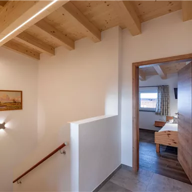 Ein heller Flur mit Holztreppe und schlichtem Design. Im Hintergrund ist ein Zimmer mit einem Fenster und Holzmöbeln sichtbar.